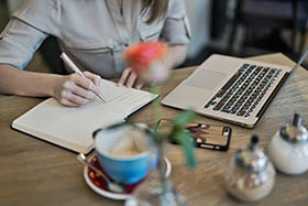femme qui écrit dans son agenda à côté d'un ordinateur d'un smartphone et d'un café